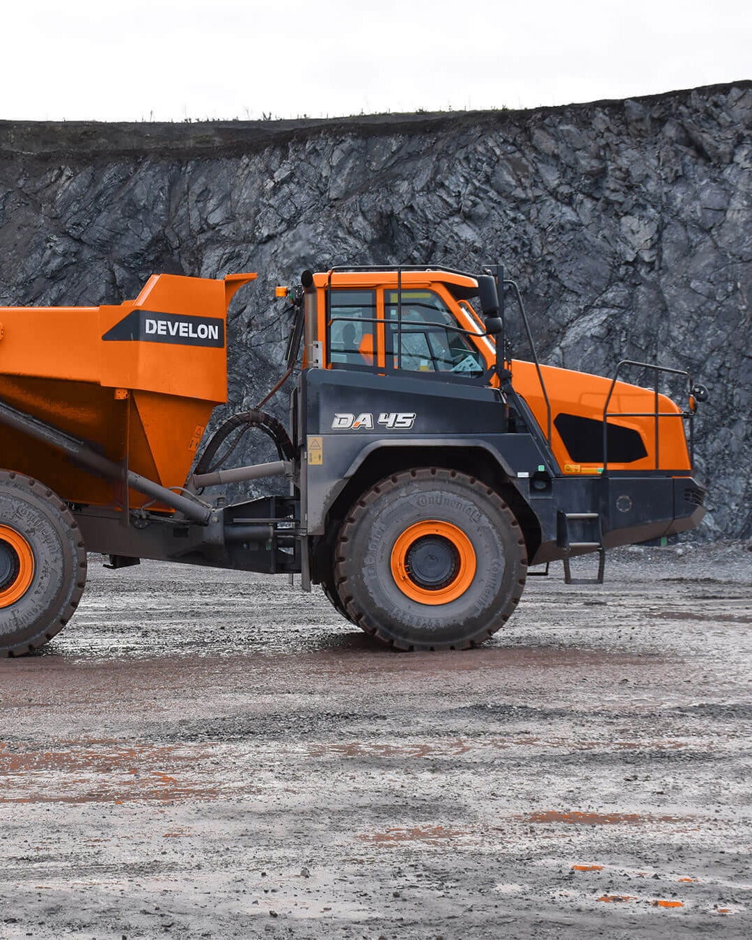A Develon DA45 Articulated Dump Truck parked on a gravel worksite, with a steep rocky cliff face in the background under an overcast sky.