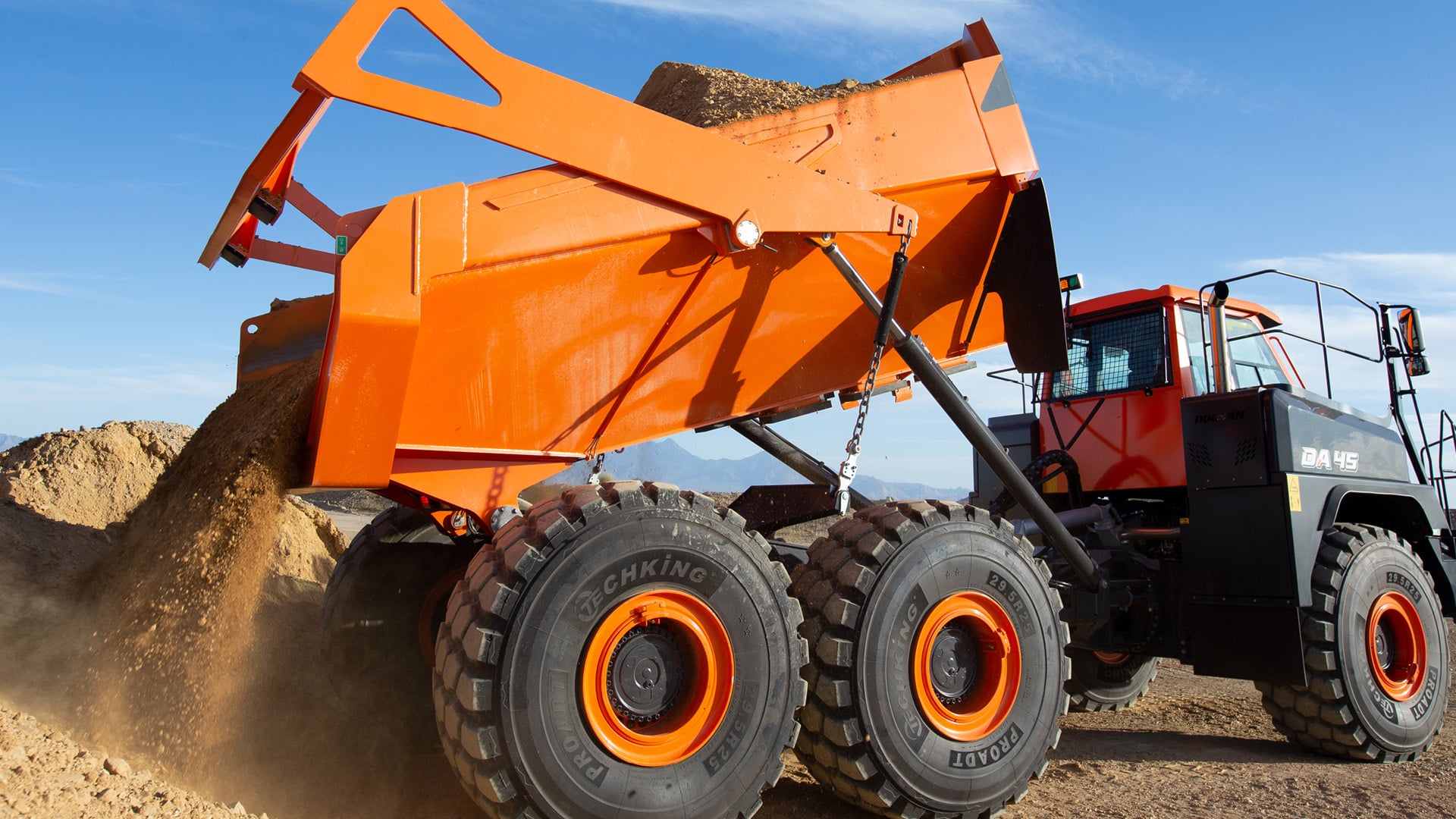A Develon DA45 Articulated Dump Truck actively unloading a full load of earth onto a construction site, with clear skies and mountains in the distance.