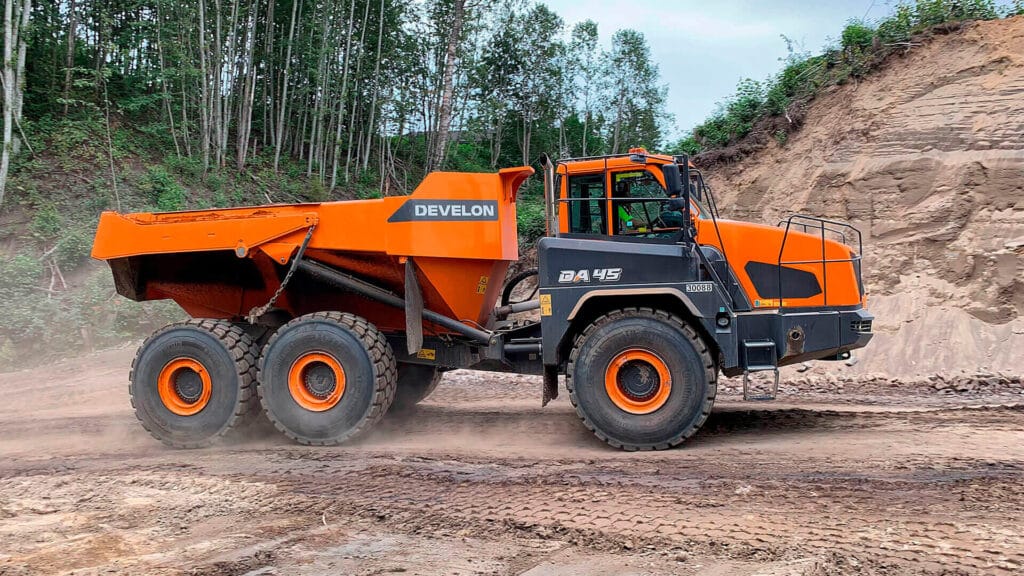 A Develon DA45 Articulated Dump Truck driving across a rugged dirt construction site, with forest trees and a sand embankment in the background.