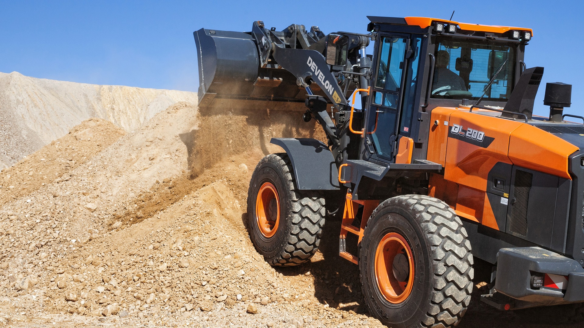 A Develon DL200A-7M Wheel Loader scooping up a pile of dirt on a sunlit construction site with rugged terrain and a clear blue sky in the background.