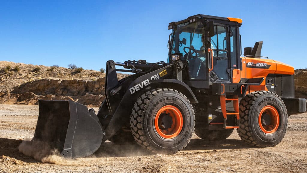 A Develon DL200A-7M Wheel Loader operating on a dry, rocky construction site under clear blue skies.