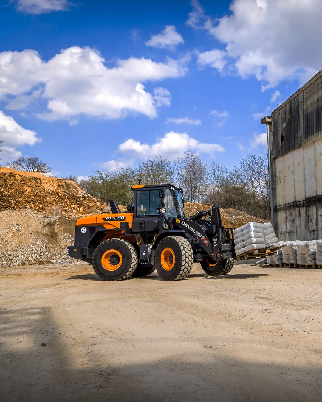 A Develon DL250-7M Wheel Loader using pallet forks to lift a load of bagged material outside an industrial site, with a clear blue sky and light cloud cover in the background.