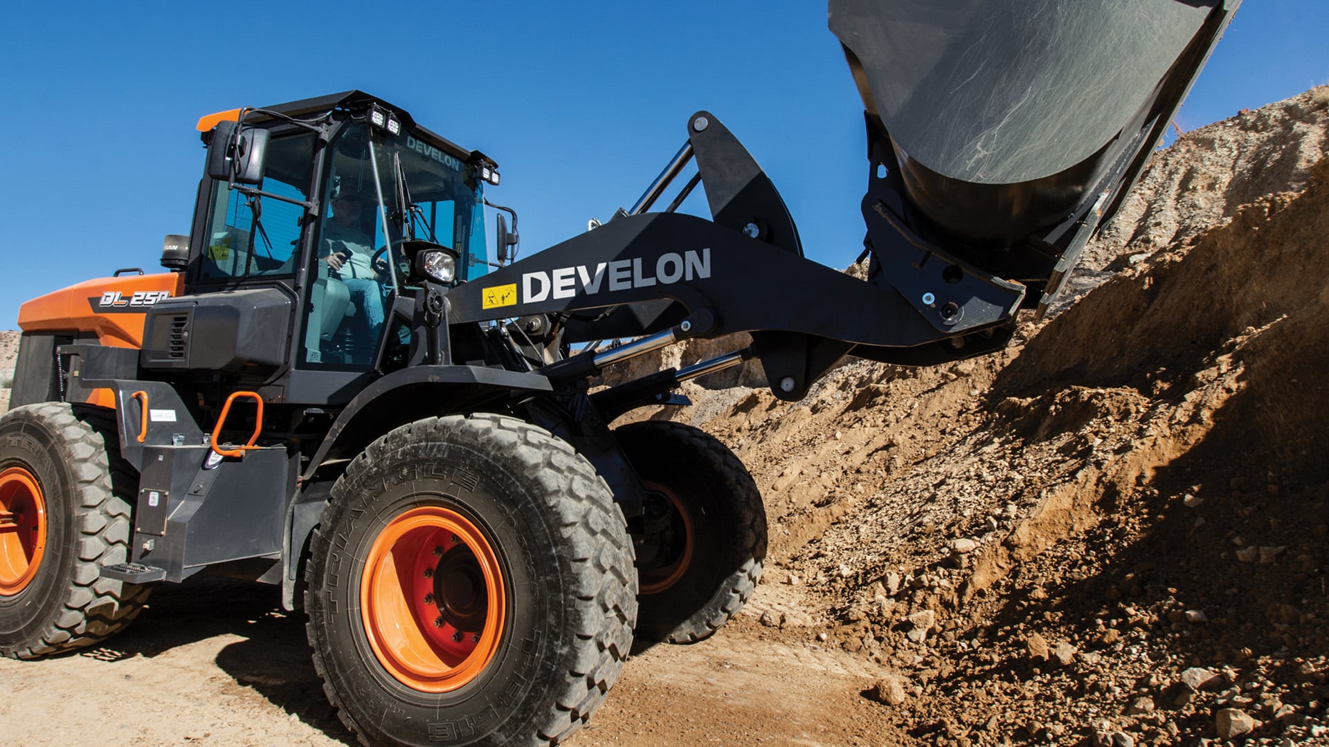 A Develon DL250-7M Wheel Loader lifting a full bucket of earth against a steep dirt slope in a quarry, with the operator clearly visible through the cab under a clear blue sky.
