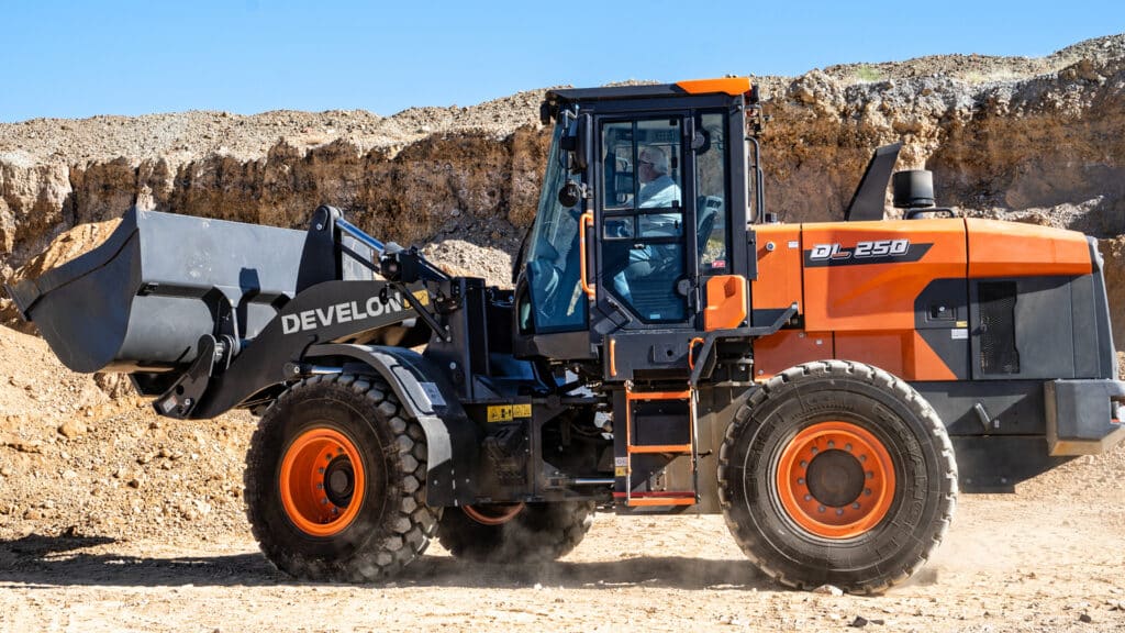 A Develon DL250-7M Wheel Loader working in a rugged quarry environment, with its bucket raised and filled, under bright daylight conditions and against a backdrop of exposed rock and dirt.
