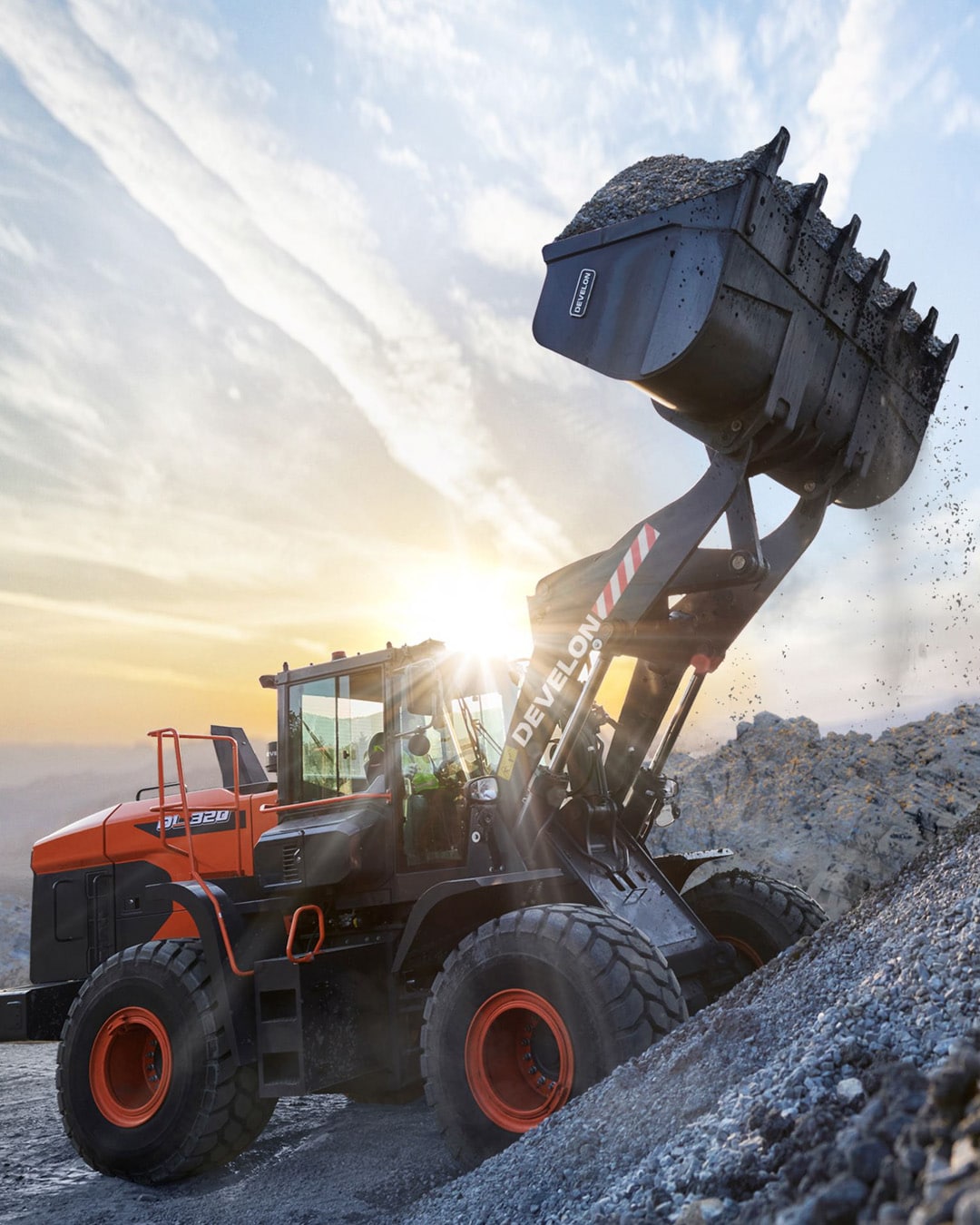 A Develon DL320-7M Wheel Loader working on a rocky slope at sunrise, lifting a full bucket of gravel against a dramatic sky backdrop.