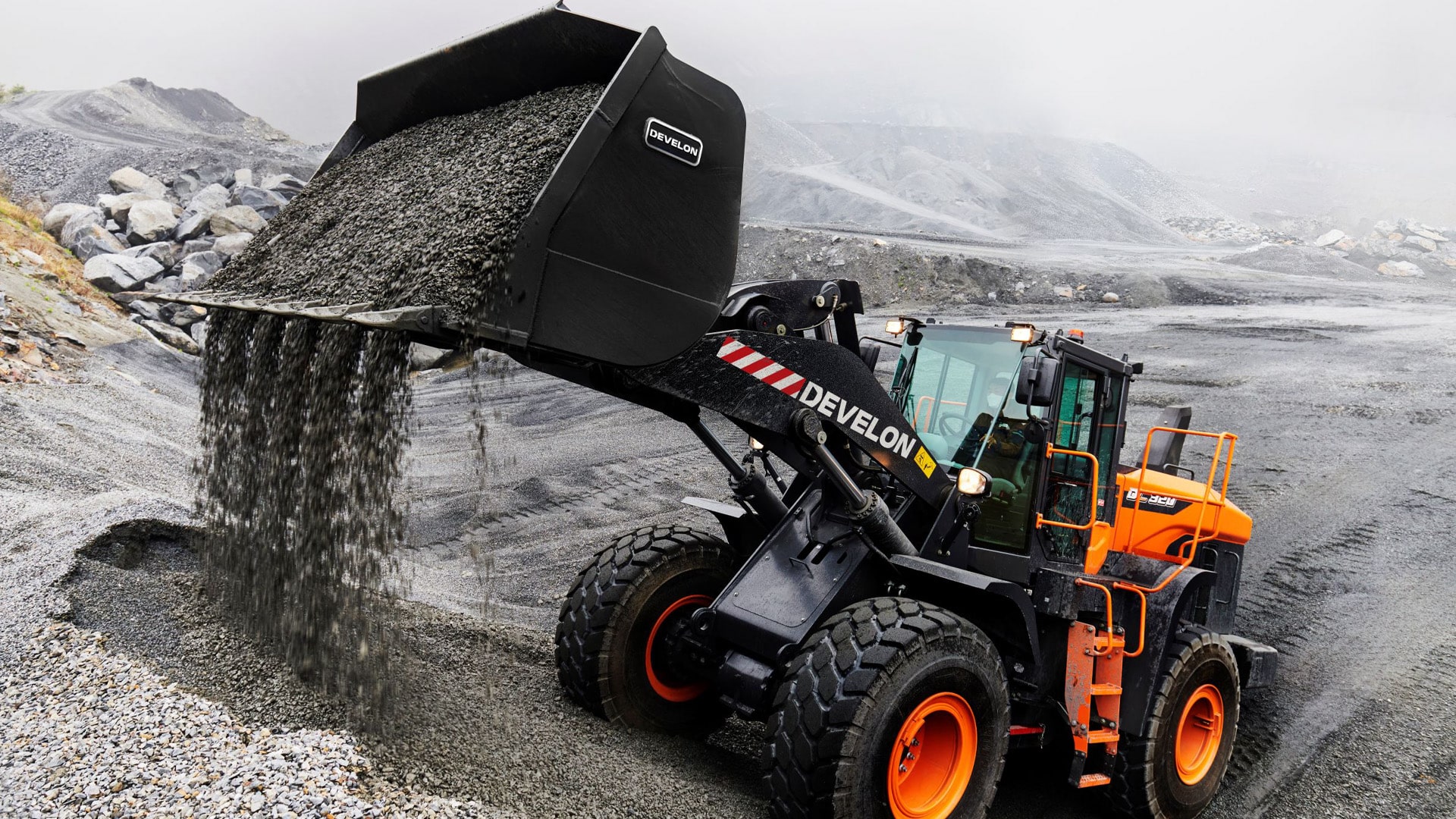 A Develon DL320-7M Wheel Loader unloading gravel at a quarry site, operating on wet terrain with foggy hills in the background.