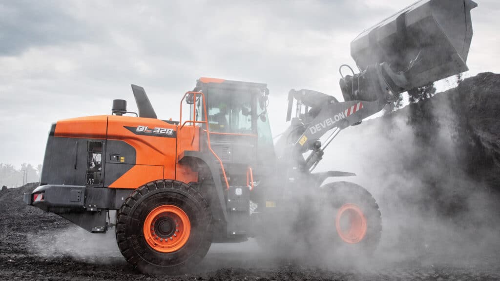 A Develon DL320-7M Wheel Loader working in wet, muddy conditions, dumping a load of material in a rugged outdoor environment under overcast skies.
