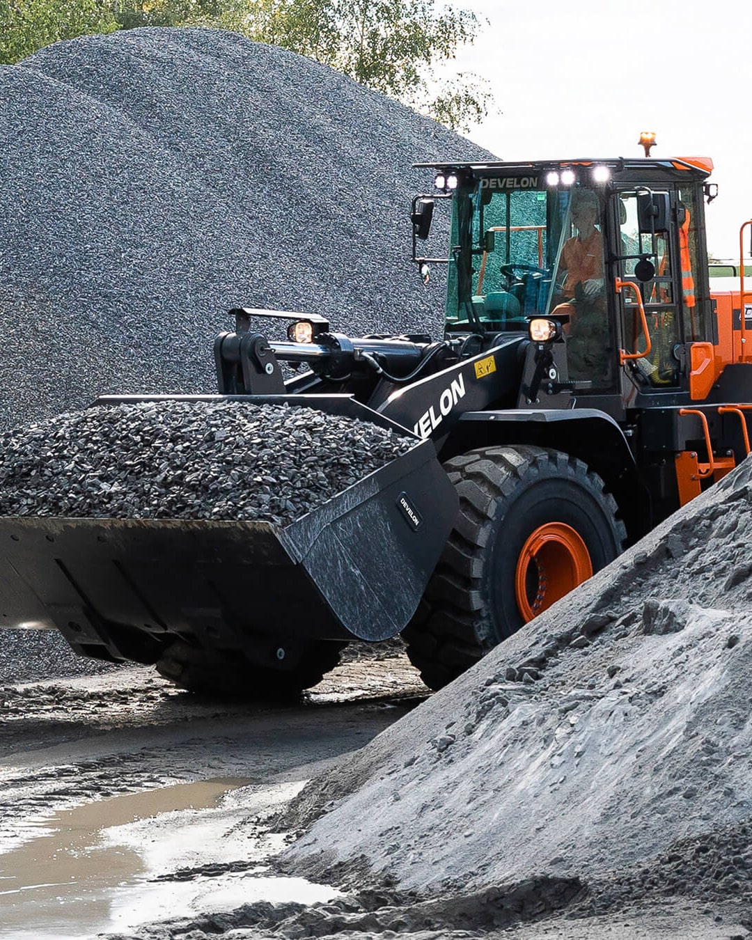 A Develon DL420-7M Wheel Loader transporting a full bucket of gravel at a quarry site, with large stockpiles of crushed stone in the background and muddy ground underfoot.