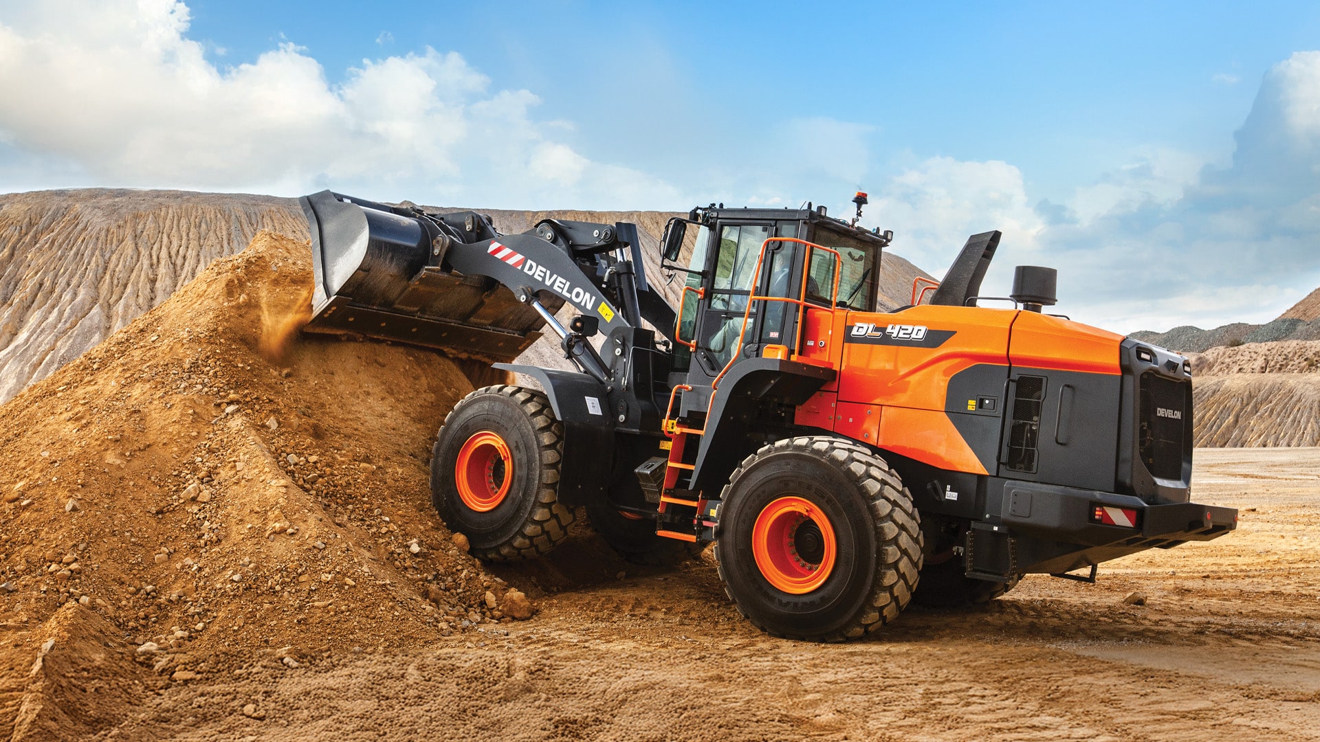 A Develon DL420-7M Wheel Loader working on a dirt mound at a construction site, lifting soil with its front bucket on rugged terrain under a bright blue sky.
