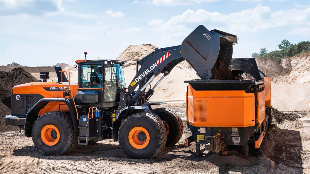 A Develon DL420-7M Wheel Loader loading material into a bright orange machine on a sandy worksite, surrounded by dirt piles and clear blue skies.