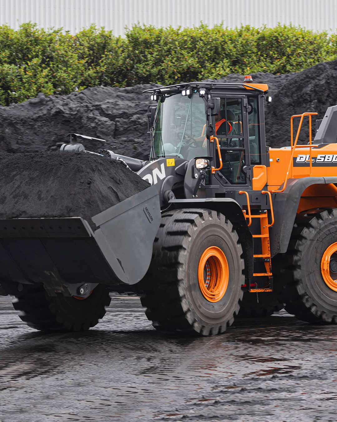 A Develon DL580-7M Wheel Loader transporting a full bucket of dark material across a wet, industrial worksite, with piles of black aggregate and green shrubs in the background.