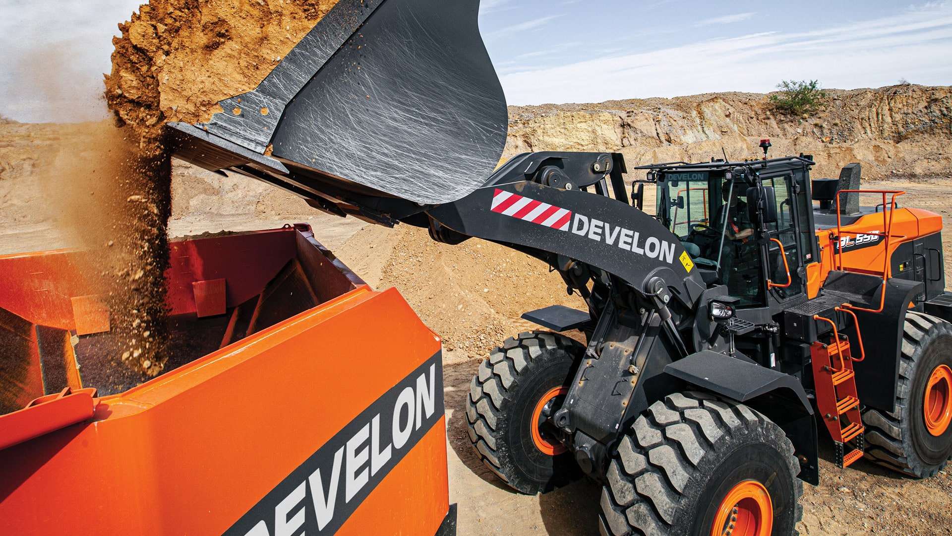 A Develon DL580-7M Wheel Loader unloading a full bucket of soil into an orange screening machine on a dusty construction site.
