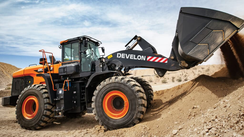 A Develon DL580-7M Wheel Loader in action, dumping a full bucket of soil on a rugged construction site under a partly cloudy sky.