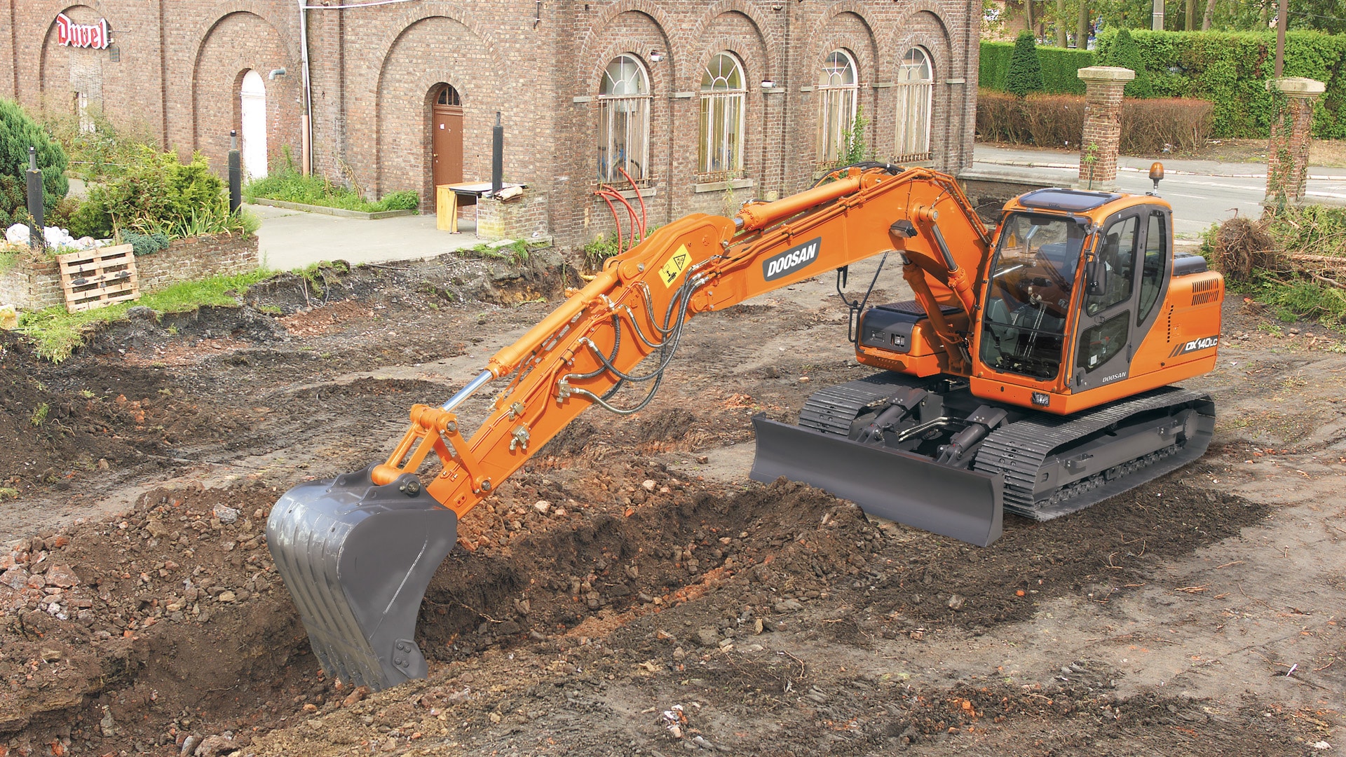 A bright orange Develon DX140LC Large Excavator operating on a construction site, digging a trench with its heavy-duty bucket. The excavator features durable rubber tracks, a spacious enclosed cabin, and a large front blade for leveling. A historic brick building with arched windows is visible in the background, surrounded by greenery and landscaping elements.