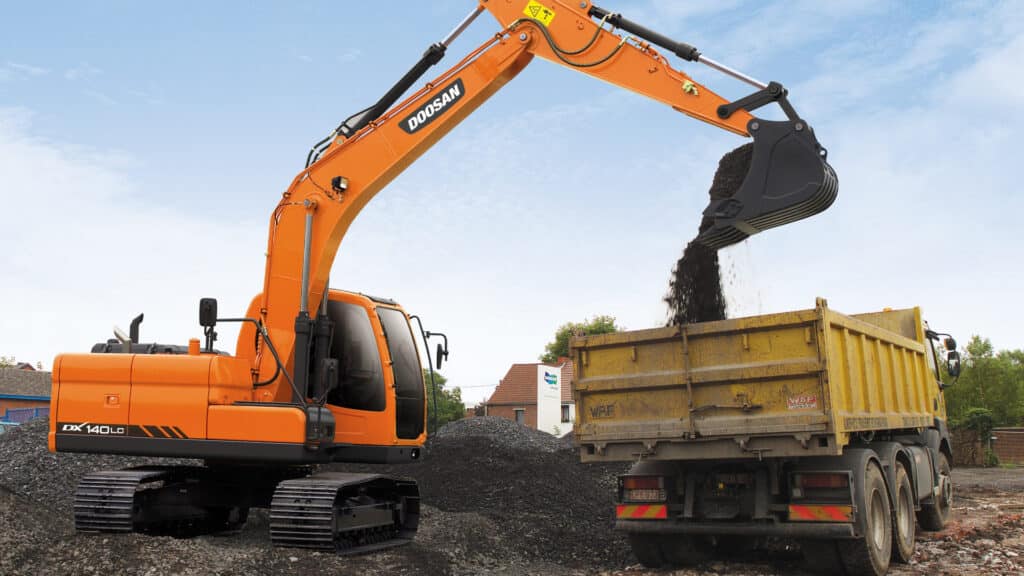 The bright orange Develon DX140LC Large Excavator efficiently loading gravel into a yellow dump truck on a construction site. The powerful arm and bucket showcase smooth hydraulic motion as the excavator transfers material with precision. The durable rubber tracks provide stability on the rocky terrain, while the modern, enclosed cab offers optimal visibility for the operator. The clear blue sky and surrounding buildings create a dynamic backdrop, emphasizing the machine’s capability in heavy-duty loading and earthmoving tasks.