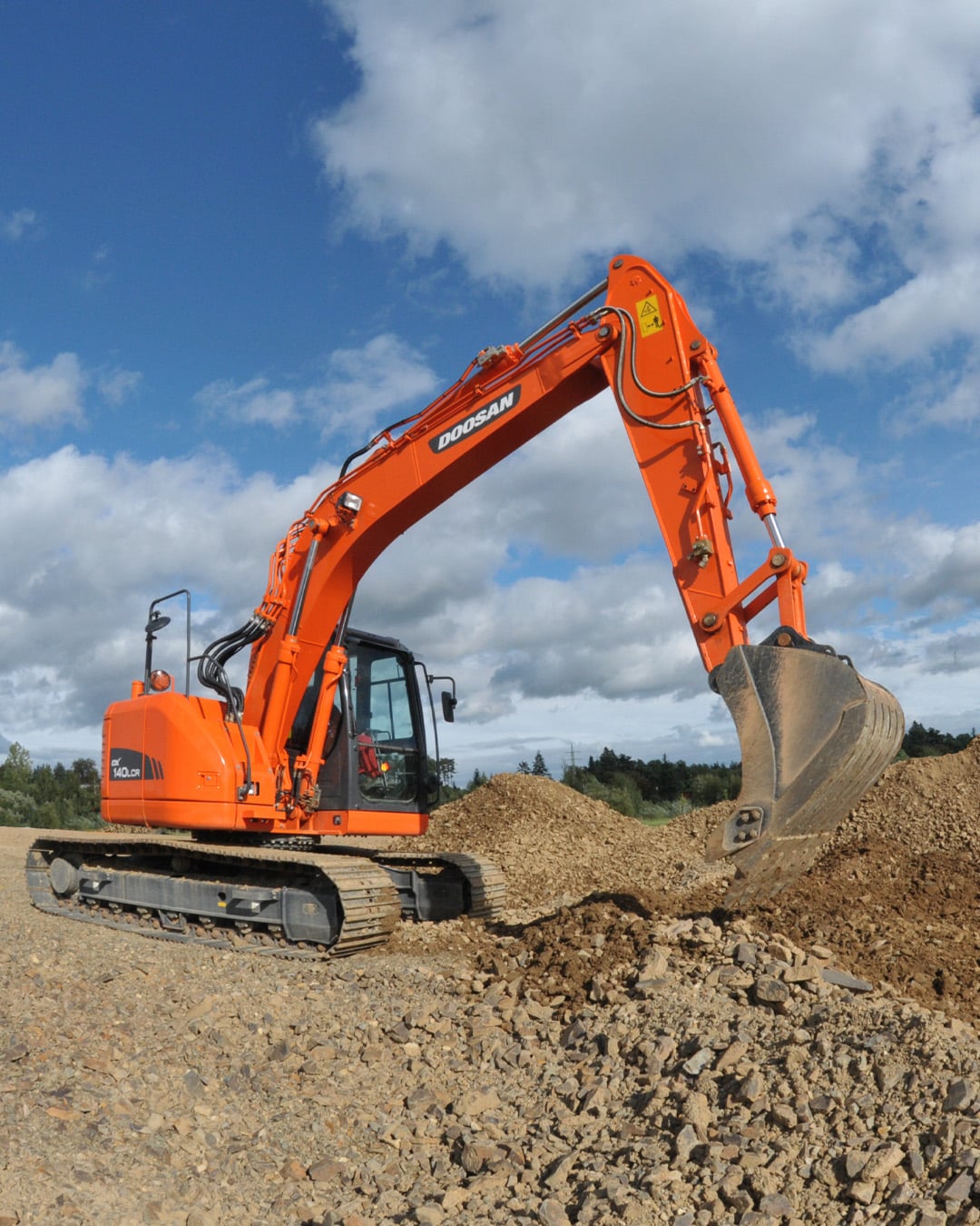 The bright orange Develon DX140LCR Large Excavator working on a rugged construction site, scooping and moving piles of gravel and dirt with its heavy-duty bucket. The excavator features durable rubber tracks, a spacious enclosed cabin with clear visibility, and robust hydraulic systems. The vibrant orange colour stands out against the clear blue sky with scattered white clouds, emphasising the machine’s power and reliability in tough terrain and earthmoving tasks.