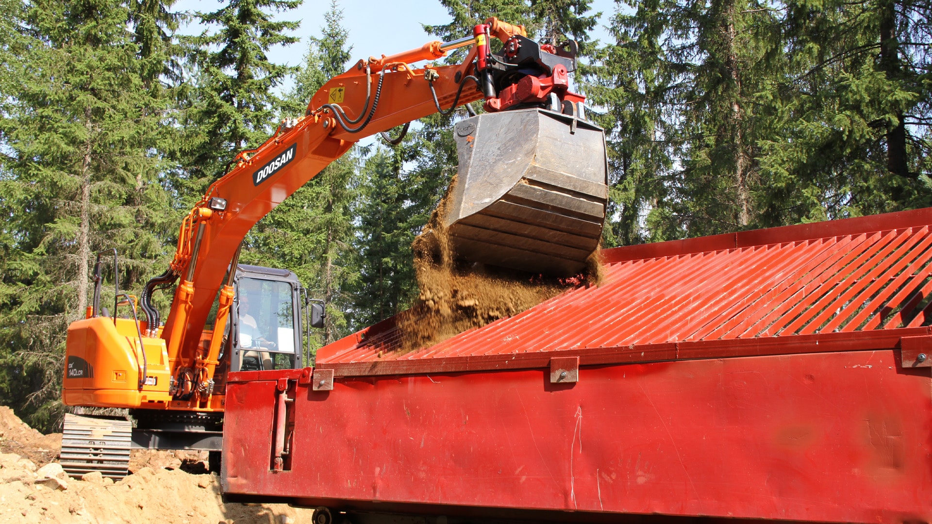 The bright orange Develon DX140LCR Large Excavator efficiently loading soil and debris into a large red screening hopper on a forestry worksite. The excavator’s robust arm and heavy-duty bucket are in action, delivering precise material placement. The spacious enclosed cabin ensures operator comfort and visibility, while the rugged rubber tracks provide stability on uneven terrain. Tall evergreen trees surround the site, highlighting the machine’s versatility in forestry and land clearing applications.