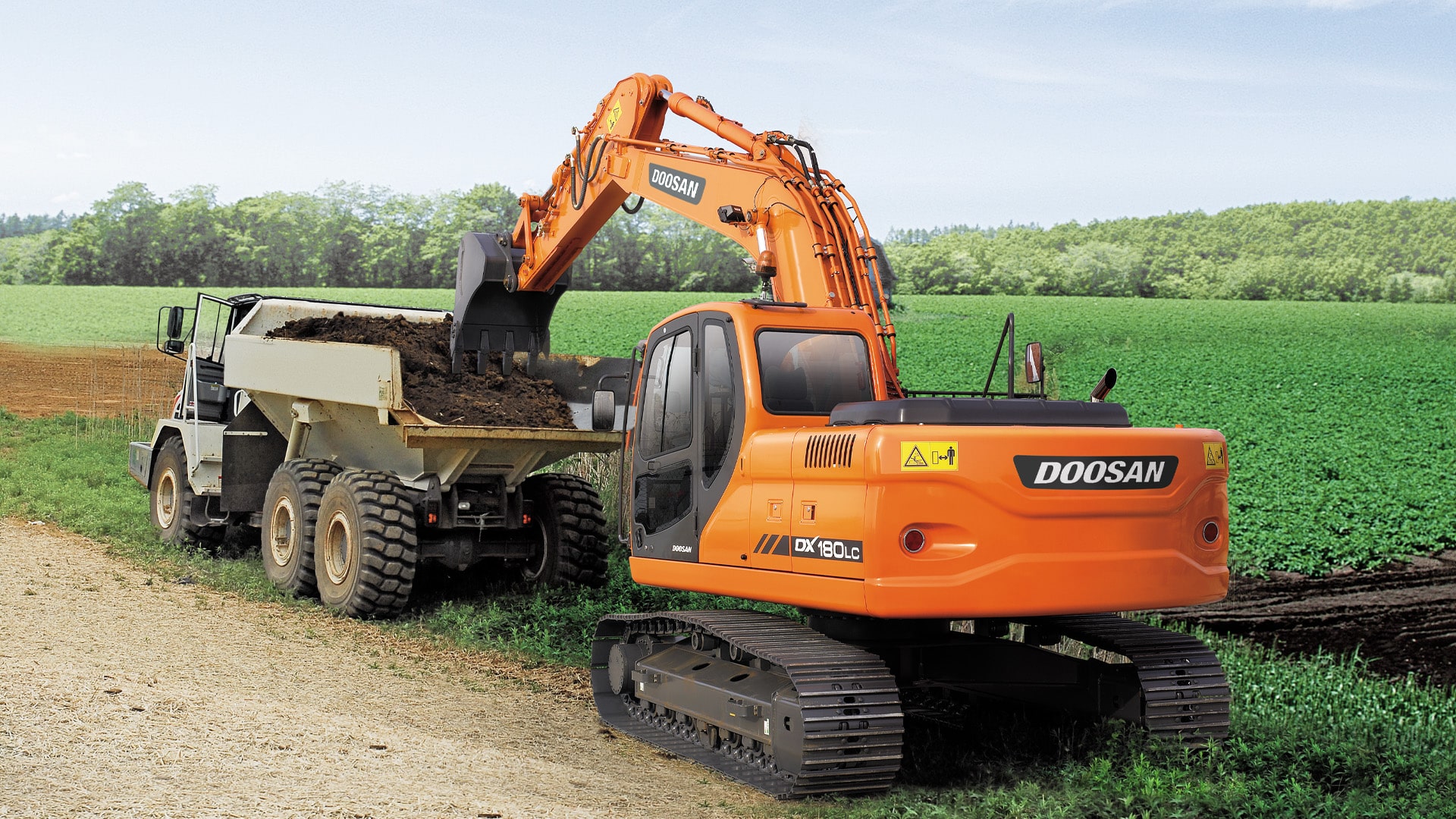 Develon DX180LCN Large Excavator loading dirt into a dump truck on a rural construction site, surrounded by lush green fields and forest in the background.