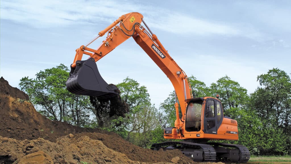 Develon DX180LCN Large Excavator scooping and dumping soil on a construction site, with lush green trees and a clear blue sky in the background.