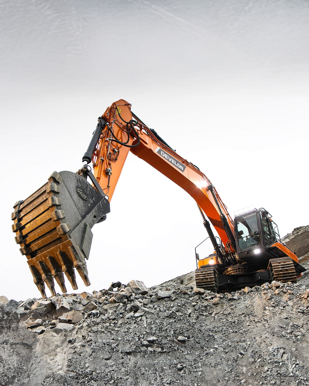 The bright orange Develon DX300LC-7M Large Excavator positioned on rugged rocky terrain, holding a massive bucket filled with crushed stone and debris. The excavator’s robust hydraulic arm and heavy-duty bucket highlight its powerful performance in demanding excavation and quarrying tasks. The spacious enclosed cabin offers optimal visibility and comfort for the operator. The durable rubber tracks ensure stability on uneven and challenging surfaces. The stark white sky background contrasts with the machine's vivid orange colour, emphasising its dominance and efficiency on the job site.