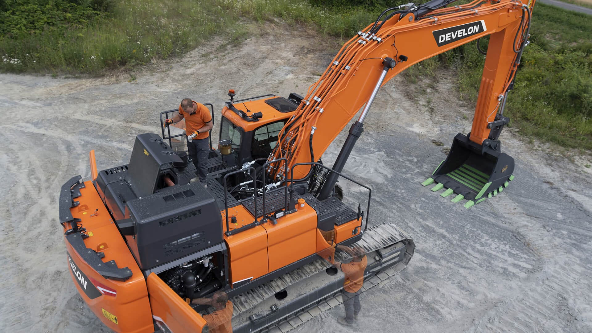 The bright orange Develon DX300LC-7M Large Excavator seen from an elevated perspective, with a worker performing maintenance on the top platform. The excavator features durable rubber tracks, a spacious enclosed cabin, and a powerful hydraulic arm with a heavy-duty bucket fitted with green wear-resistant teeth. The machine’s robust design and easy-access maintenance areas highlight its efficiency and operator convenience. The surrounding dirt and grassy landscape emphasise the excavator’s rugged adaptability in various terrain conditions.