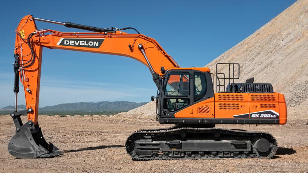 The bright orange Develon DX300LC-7M Large Excavator positioned on a flat, open construction site with a backdrop of rugged mountains and a clear blue sky. The excavator features durable rubber tracks, a spacious enclosed cabin with large windows, and a powerful hydraulic arm equipped with a heavy-duty digging bucket. The sleek design and robust build emphasize the machine’s efficiency and reliability for heavy-duty earthmoving tasks. The vibrant orange colour stands out against the natural landscape, highlighting its presence and capability on demanding job sites.