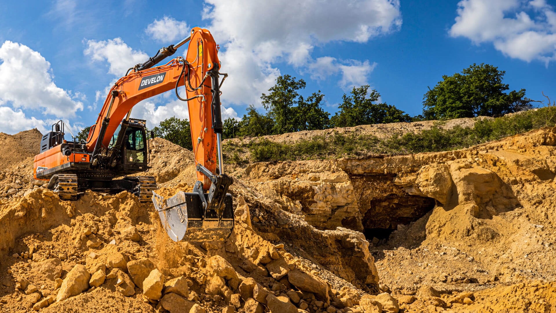The powerful Develon DX400LC-7M Large Excavator at a rugged excavation site, expertly maneuvering rocky terrain with its robust bucket and heavy-duty tracks. The vivid orange exterior contrasts against the natural rock landscape, highlighting its durability and superior performance in challenging environments.