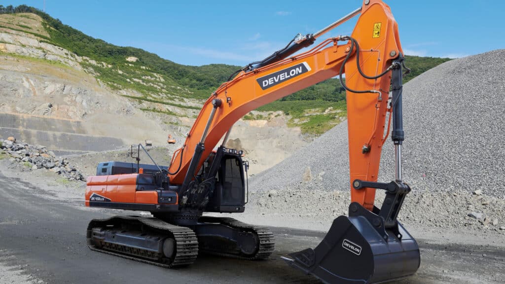 The powerful Develon DX400LC-7M Large Excavator positioned at a gravel quarry, showcasing its robust arm and heavy-duty bucket against a backdrop of rugged terrain and green hills. The bright orange finish and precise hydraulic components highlight its engineering excellence and capability for demanding excavation tasks.