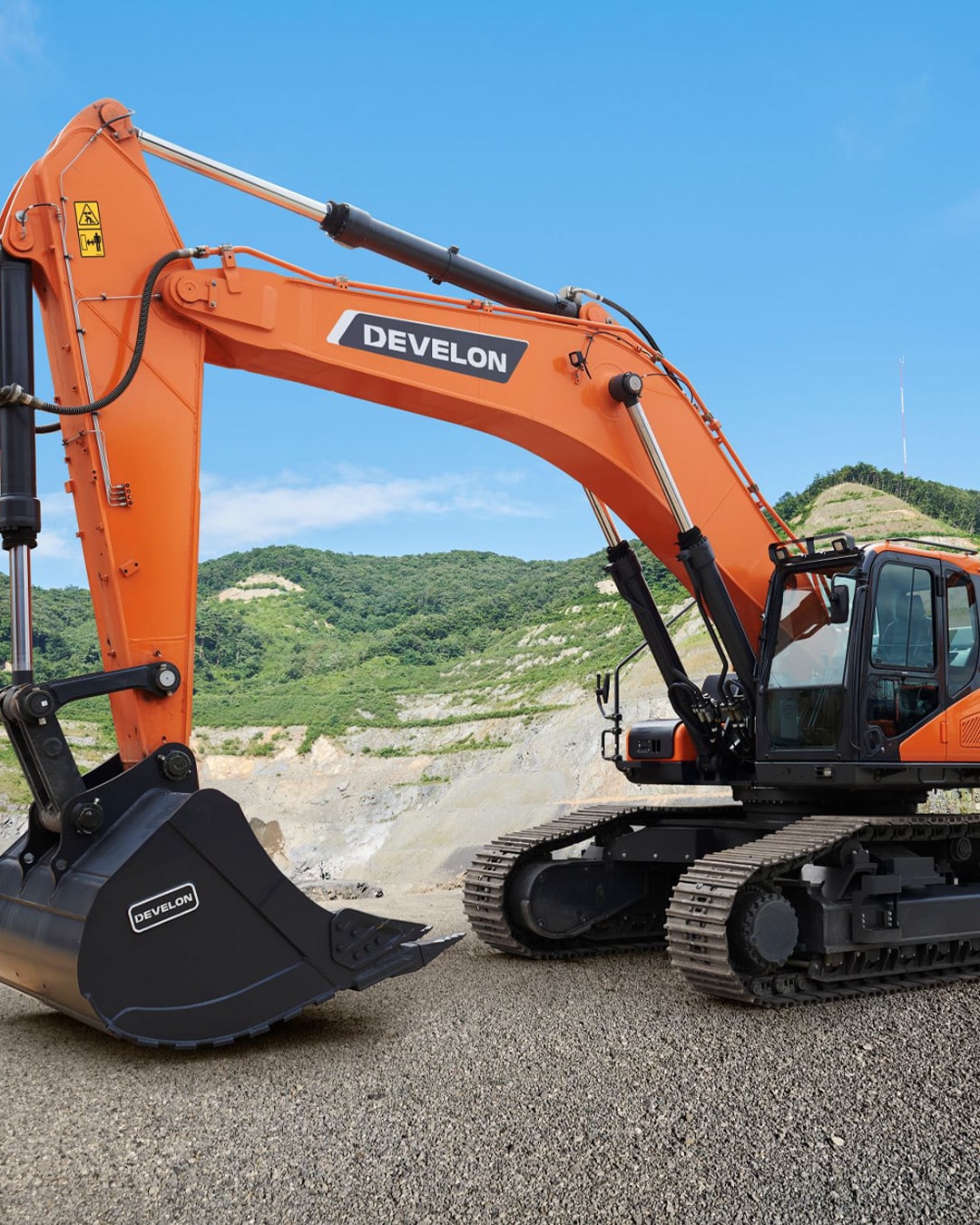 The Develon DX490LC-7M Large Excavator positioned at a quarry site, showcasing its powerful hydraulic arm and rugged bucket. The vivid orange colour contrasts against the gravel terrain and lush green hills in the background, highlighting its heavy-duty performance and durability in demanding excavation environments.