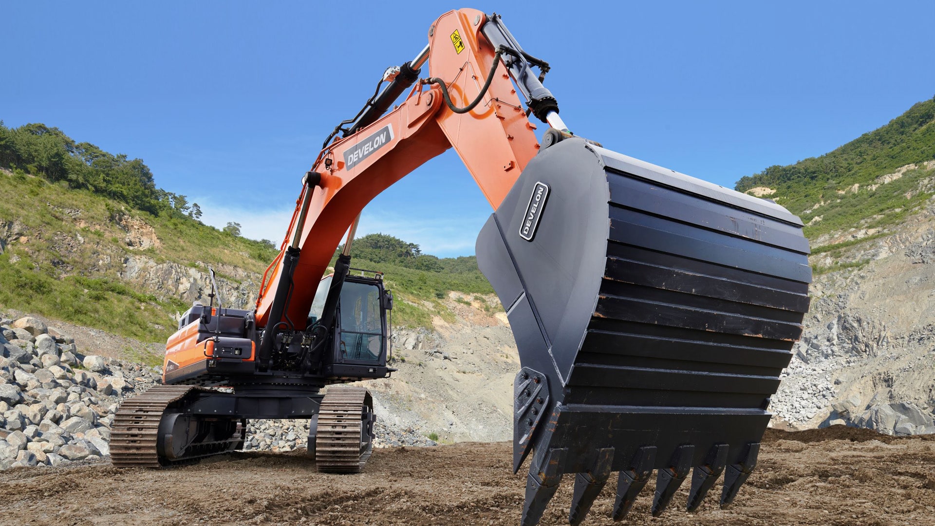 The Develon DX490LC-7M Large Excavator with a robust bucket attachment, positioned in a rugged quarry environment. The excavator's vibrant orange body contrasts against the earthy tones and rocky hillside, showcasing its power and capability in heavy-duty digging and earthmoving applications.