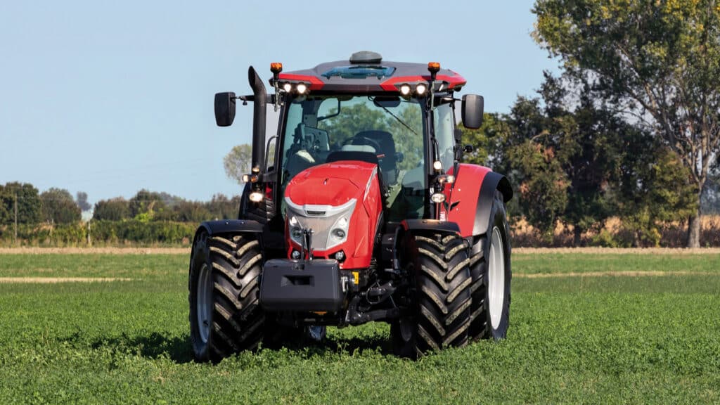 McCormick Farm Tractor driving across a lush green field, viewed from the front with trees in the background.