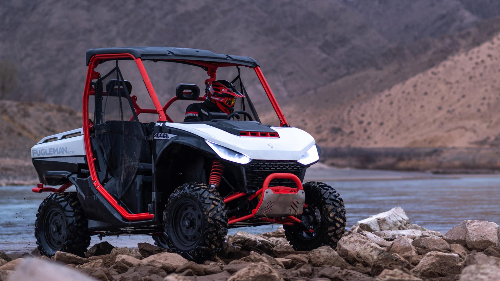 A Segway UT10 Rugged EPS UTV with LED headlights and red accents climbs over rocky terrain beside a mountain lake, driven by a rider in red gear, emphasizing its 4x4 performance and rugged off-road capability.