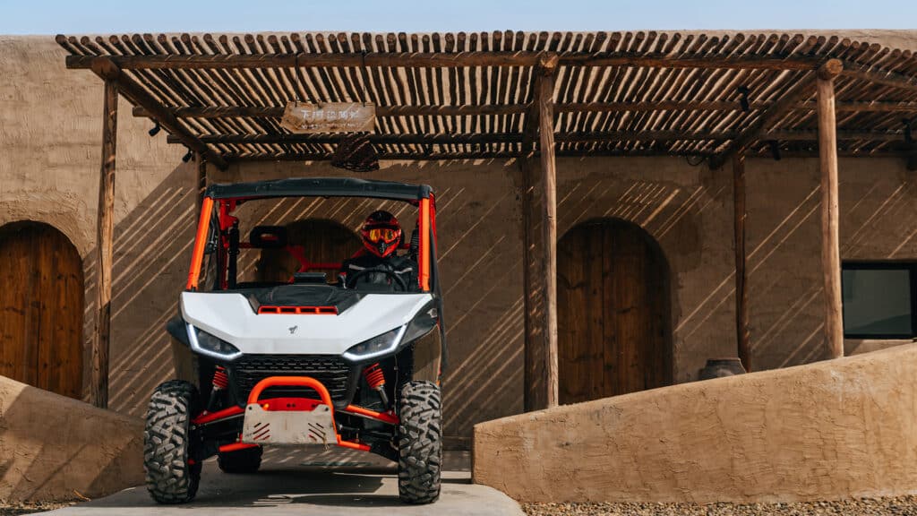A Segway UT10 Rugged EPS UTV with red accents and aggressive styling is parked under a rustic wooden pergola in front of an adobe-style building, with a rider in red gear seated behind the wheel.