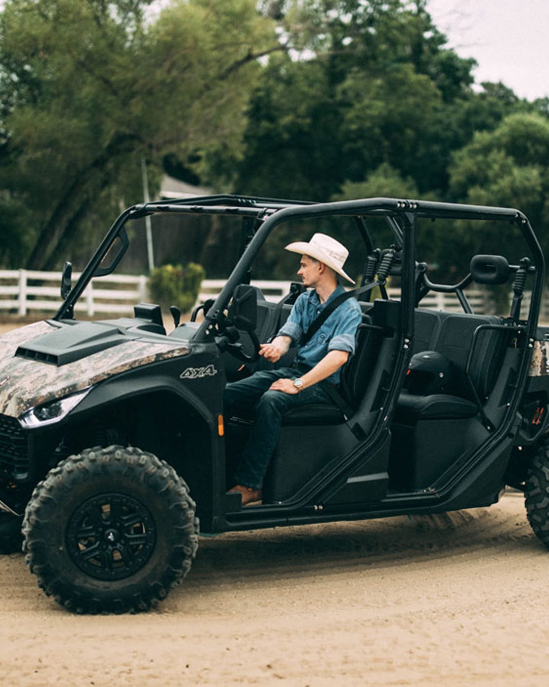A man in a cowboy hat and denim shirt sits in a Segway UT10 Crew UTV with a camo hood, parked on a dirt track near a white fence and surrounded by trees.