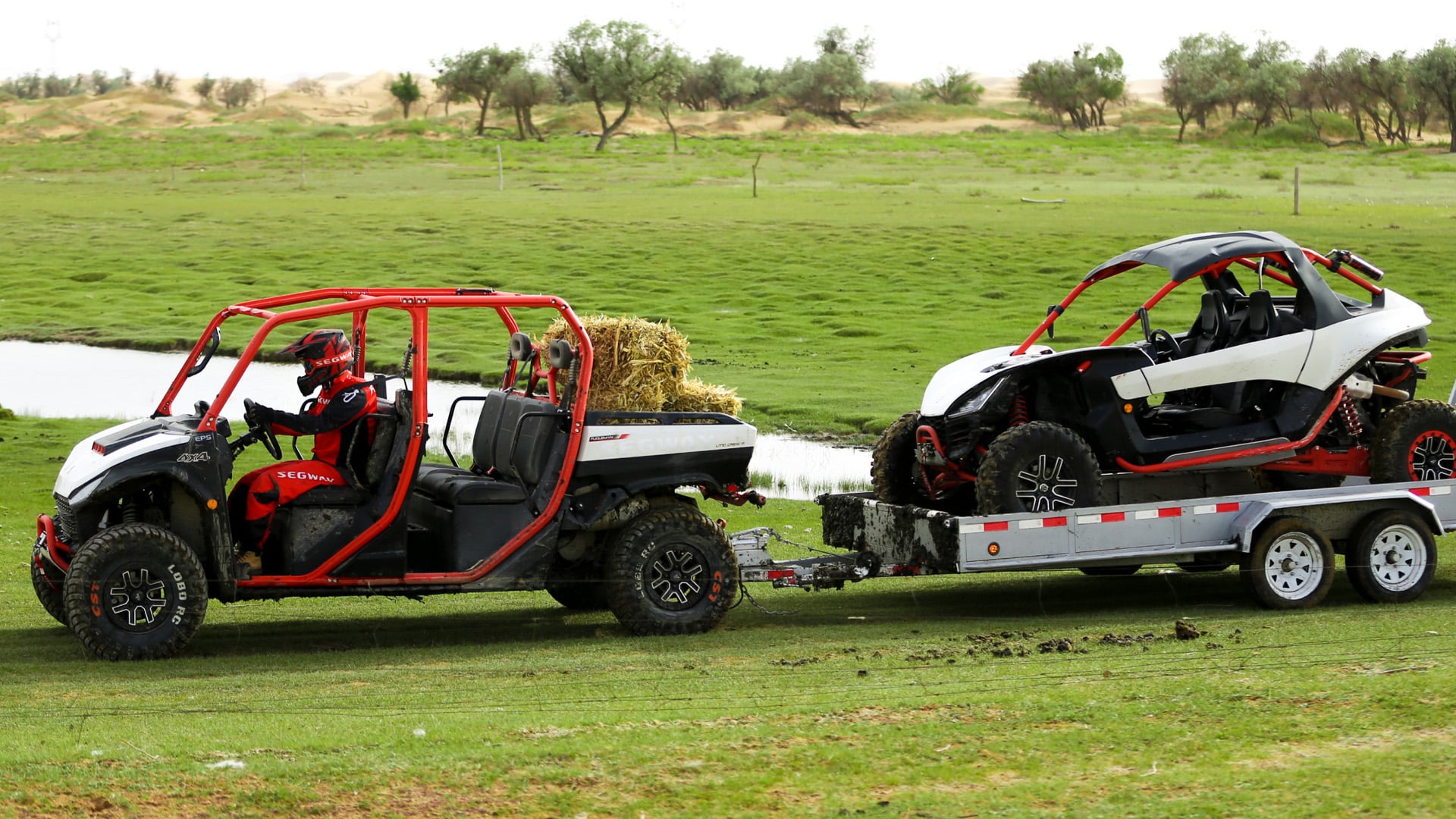 A Segway UT10 Crew UTV loaded with hay bales tows another Segway off-road vehicle on a trailer across a grassy field near a waterway, demonstrating its power and utility.