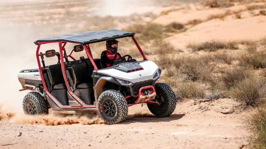 A Segway UT10 Crew UTV with a black roof and red frame drives at speed through a dusty desert track, highlighting its power, control, and all-terrain capability.