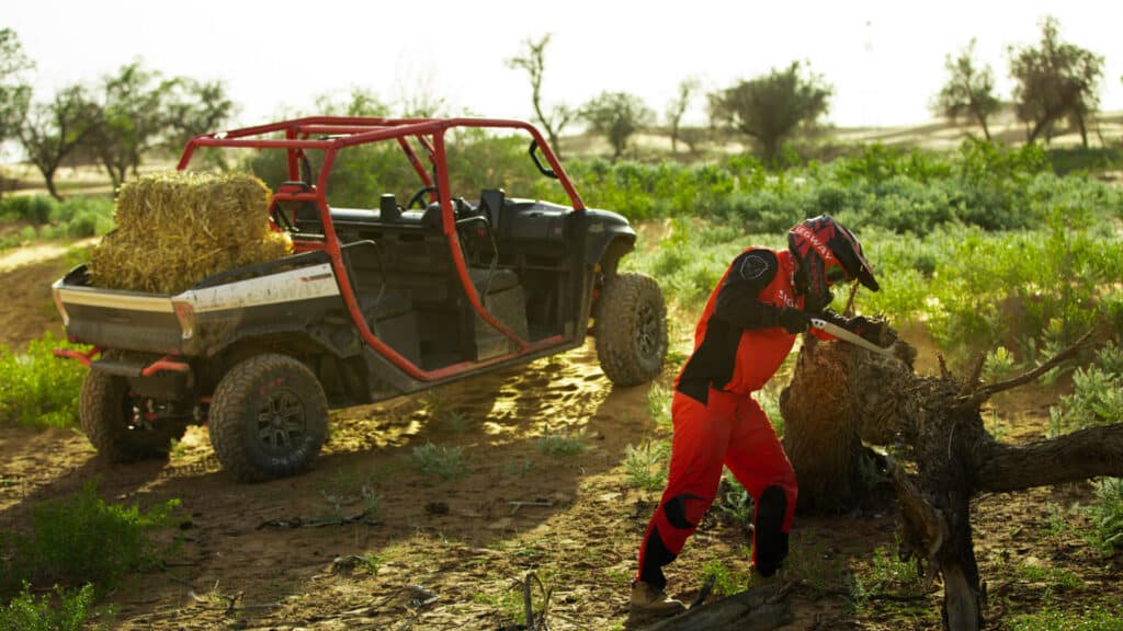 A Segway UT10 Crew Premium UTV loaded with hay bales is parked on a dirt track, while a rider in red Segway gear chops a fallen tree branch in a rugged, rural landscape.