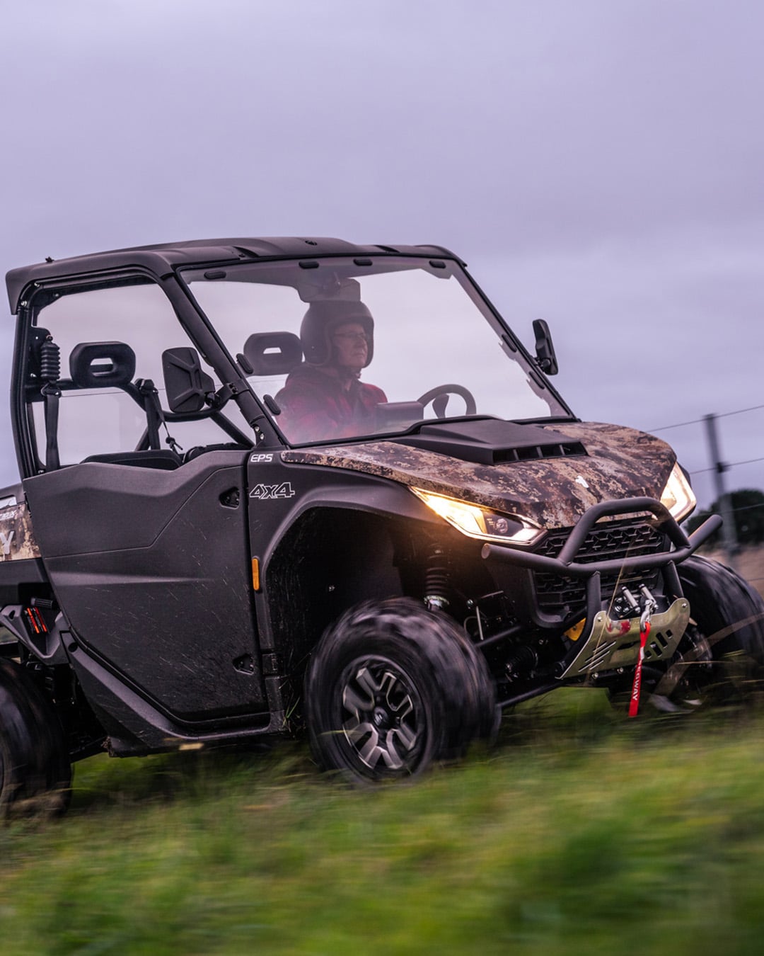 A Segway UT10 Navigator UTV with camo bodywork and headlights on drives across a grassy field at dusk, piloted by a helmeted driver, highlighting its 4x4 capability and rugged design.