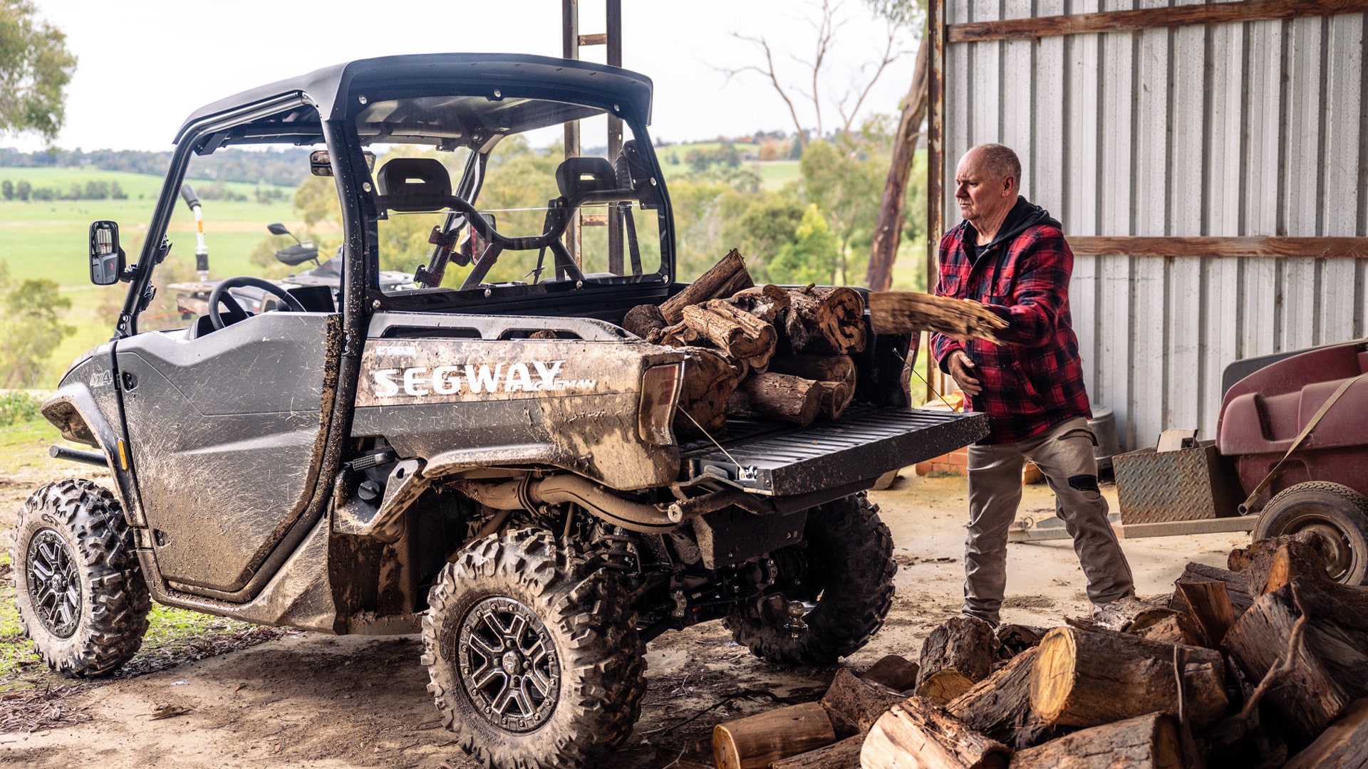 A muddy Segway UT10 Navigator UTV is parked in a shed as a man in a red flannel shirt loads firewood into the cargo bed, showcasing its durability and practicality for farm work.