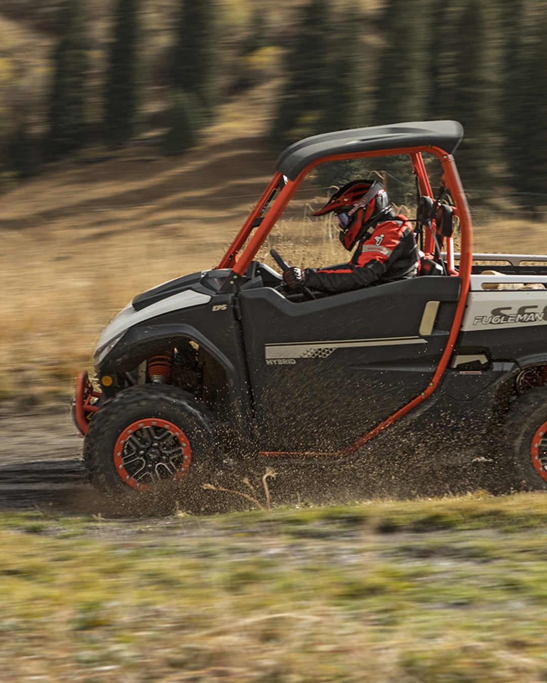 A Segway UT10 Pro UTV with hybrid power and red accents splashes through a muddy trail in a mountain meadow, driven by a rider in red gear and helmet.