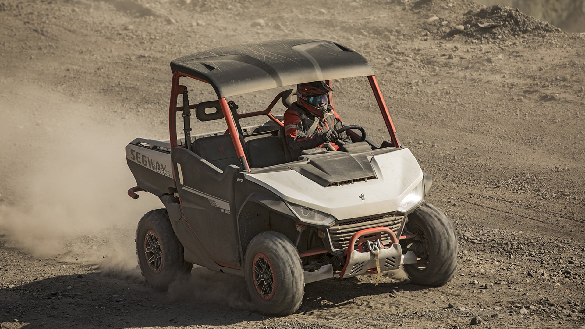 A Segway UT10 Pro UTV with a black roof and red frame kicks up dust as it navigates a rocky, rugged terrain, driven by a rider in red gear, showcasing its hybrid power and off-road endurance.