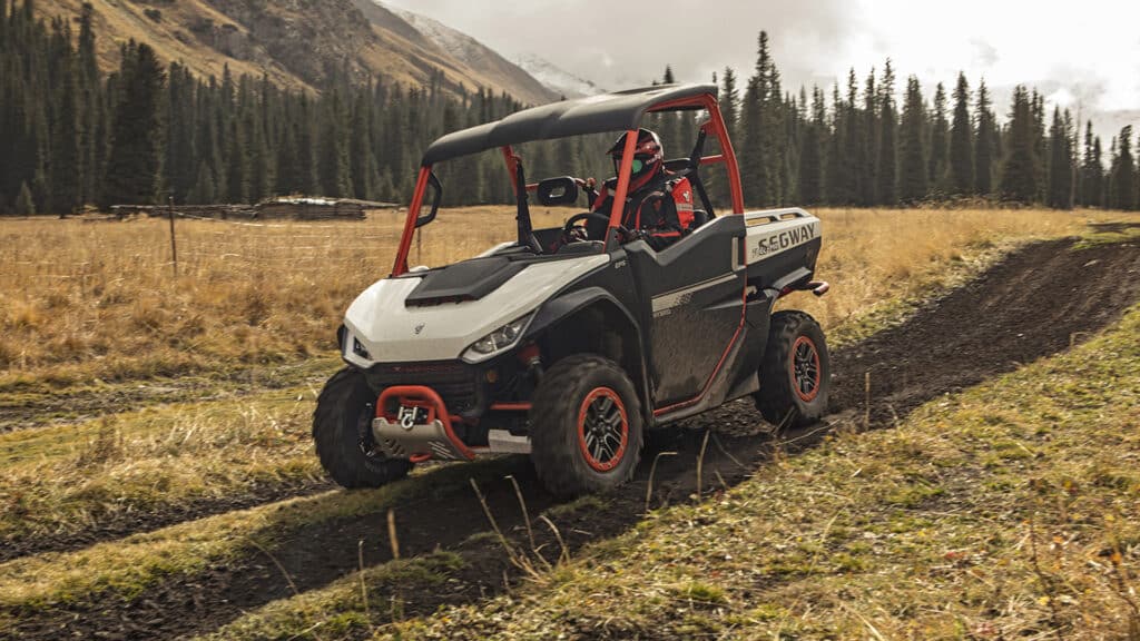 A Segway UT10 Pro UTV with red accents and a black roof drives along a muddy trail through a mountain valley, showcasing its off-road capability and rugged performance in alpine terrain.