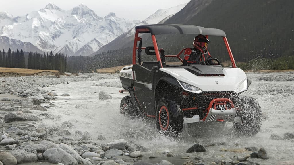 A UTV splashes through a rocky mountain stream with snow-capped peaks in the background, showcasing its off-road capabilities in rugged alpine terrain.