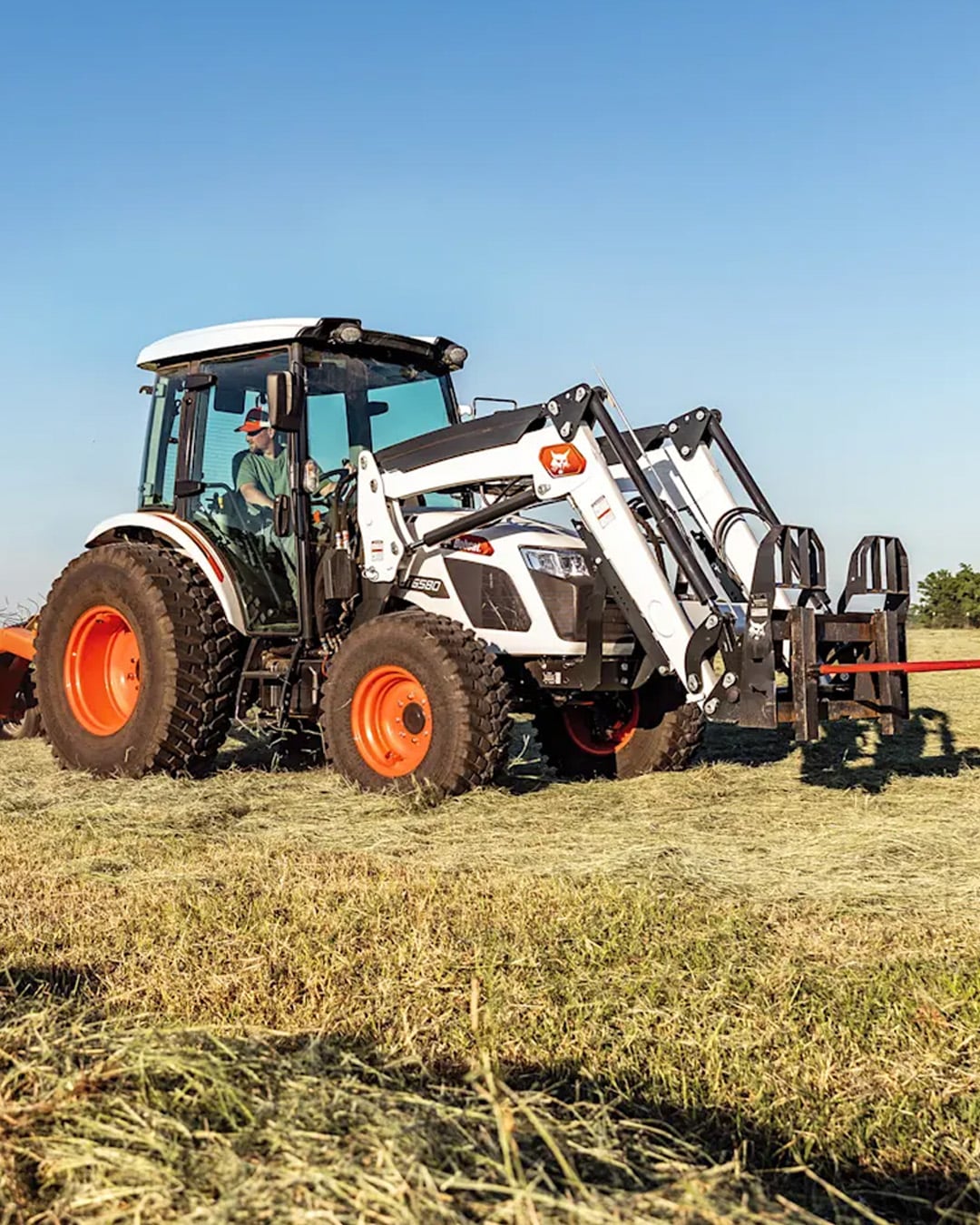 A Bobcat Utility Tractor equipped with a front-end loader and grapple attachment operates in a hay field, driven by a man inside the cab under a clear blue sky.