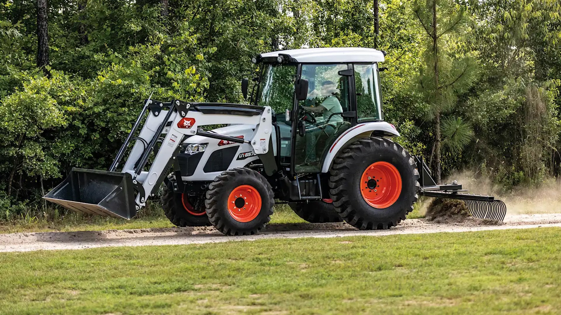 A Bobcat Utility Tractor with a front-end loader and rear landscape rake attachment drives along a dirt path beside a forested area, kicking up dust as it levels the ground.