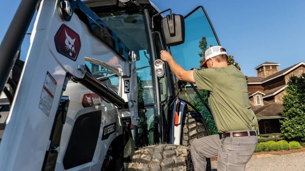 A man steps into the cab of a Bobcat Utility Tractor parked in front of a large countryside home with manicured landscaping and a clear blue sky.