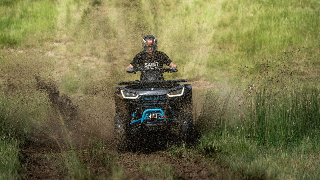 Utility vehicle powering through muddy terrain, splashing dirt while a rider in a helmet and SAINT shirt maintains control across rugged grassy landscape.
