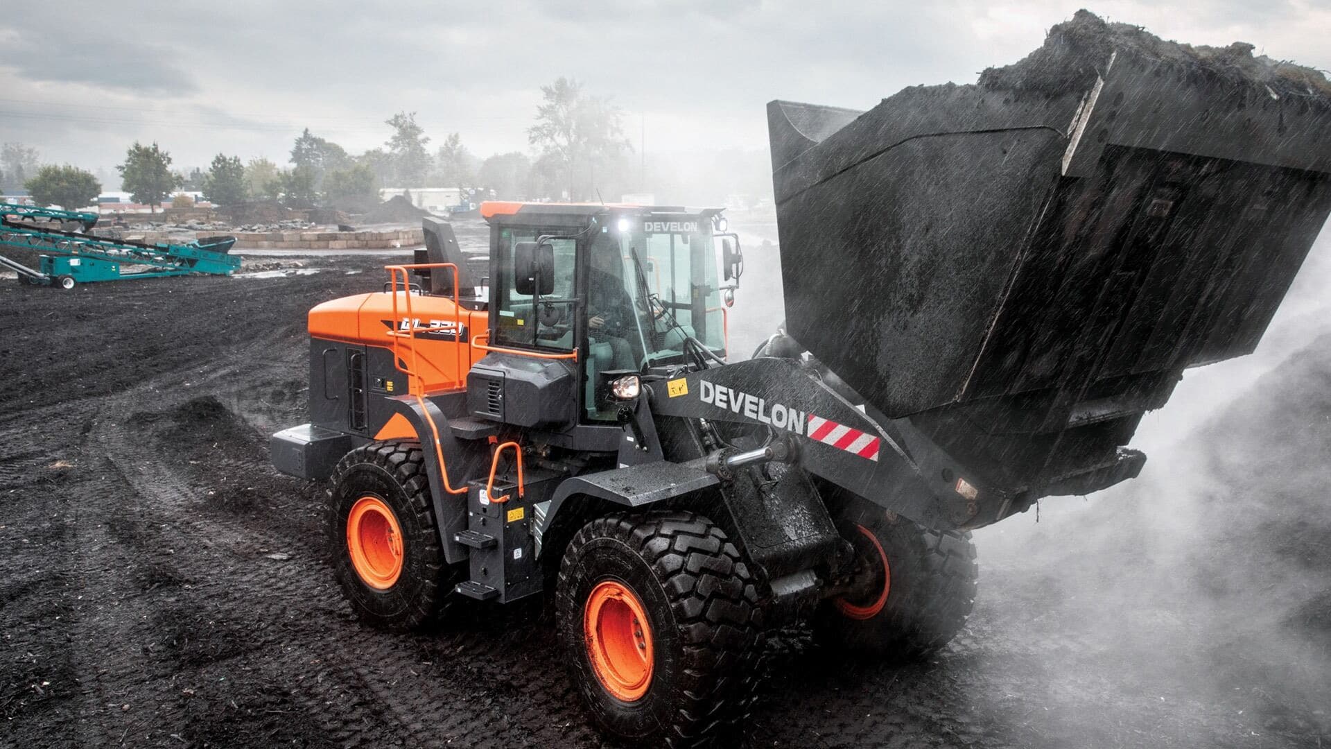 A Develon Wheel Loader working in harsh, wet conditions, efficiently moving heavy loads of material with its robust front bucket, demonstrating power and durability on a rugged job site.