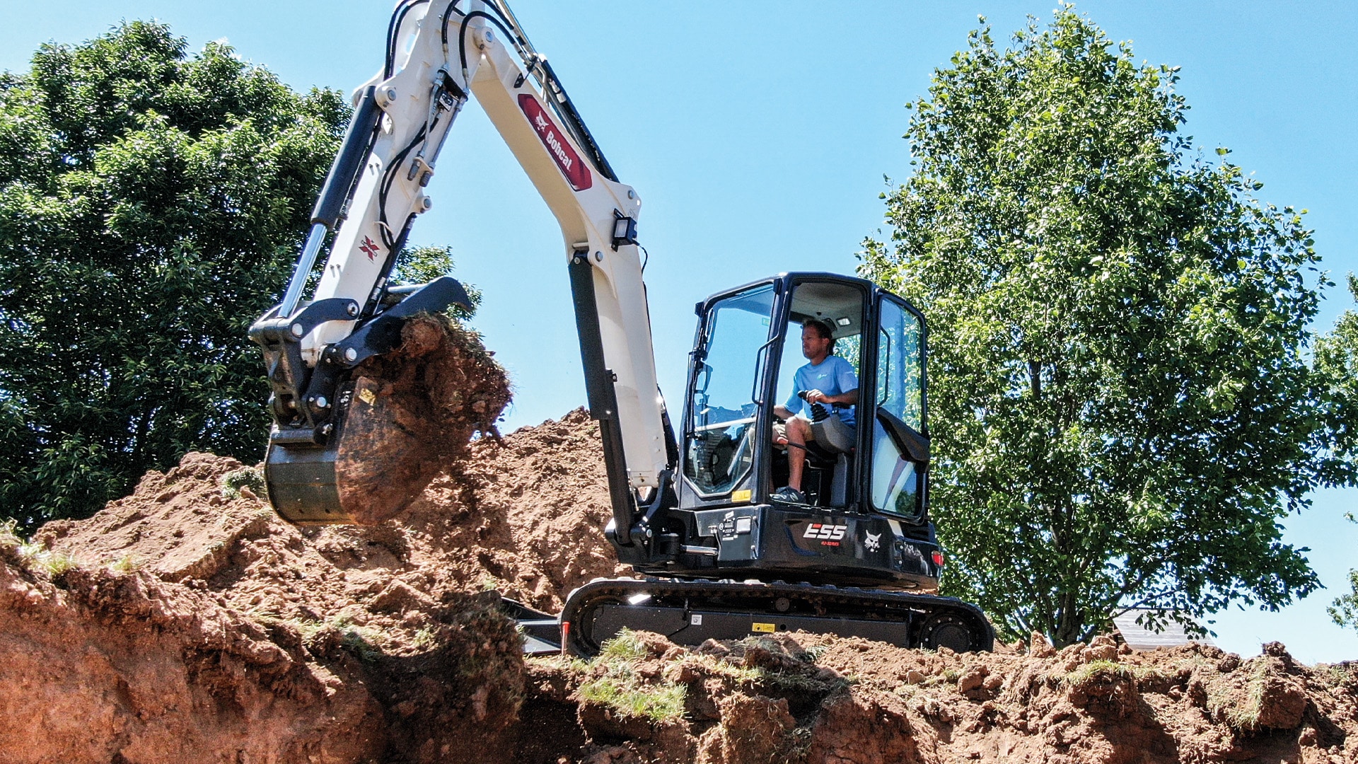 A Bobcat E55R Mini Excavator digging into a soil embankment on a sunny day, operated by a worker in a blue shirt, surrounded by lush green trees in the background.