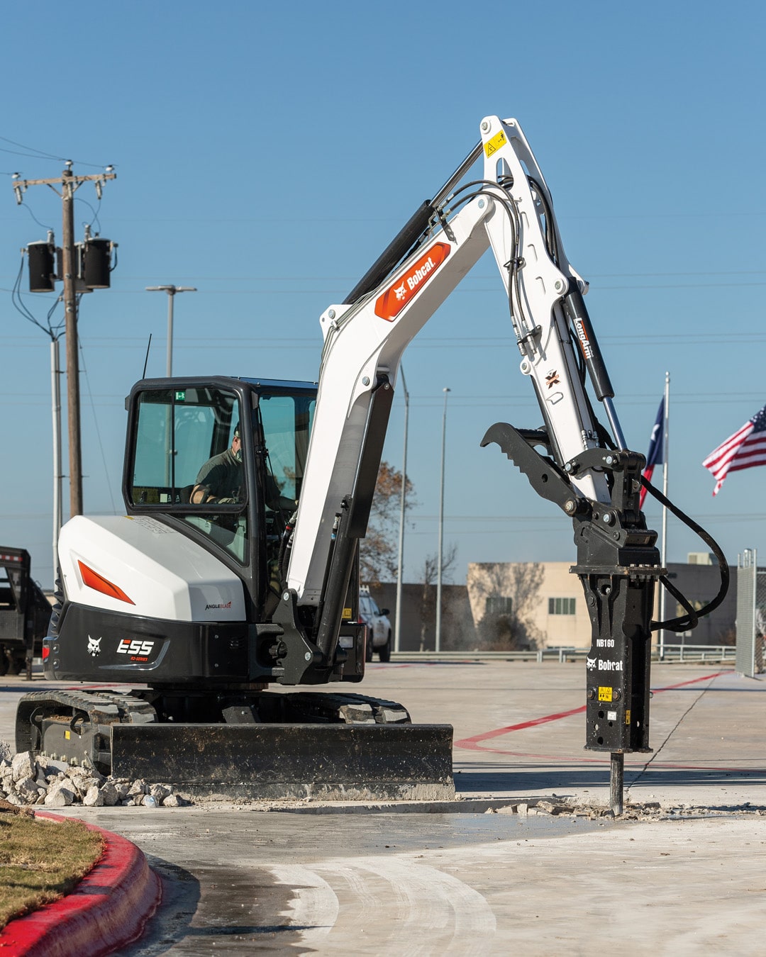 A Bobcat E55R Mini Excavator equipped with a hydraulic breaker attachment, breaking concrete on a commercial site. The excavator demonstrates power and precision, with American flags and utility poles visible in the background.